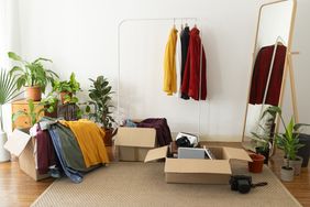 A clothing rack with hanging garments boxes with clothes and items surrounded by plants and a mirror in a room