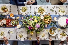 A formal dining table with plates of pasta bread flowers centerpiece surrounded by people holding glasses