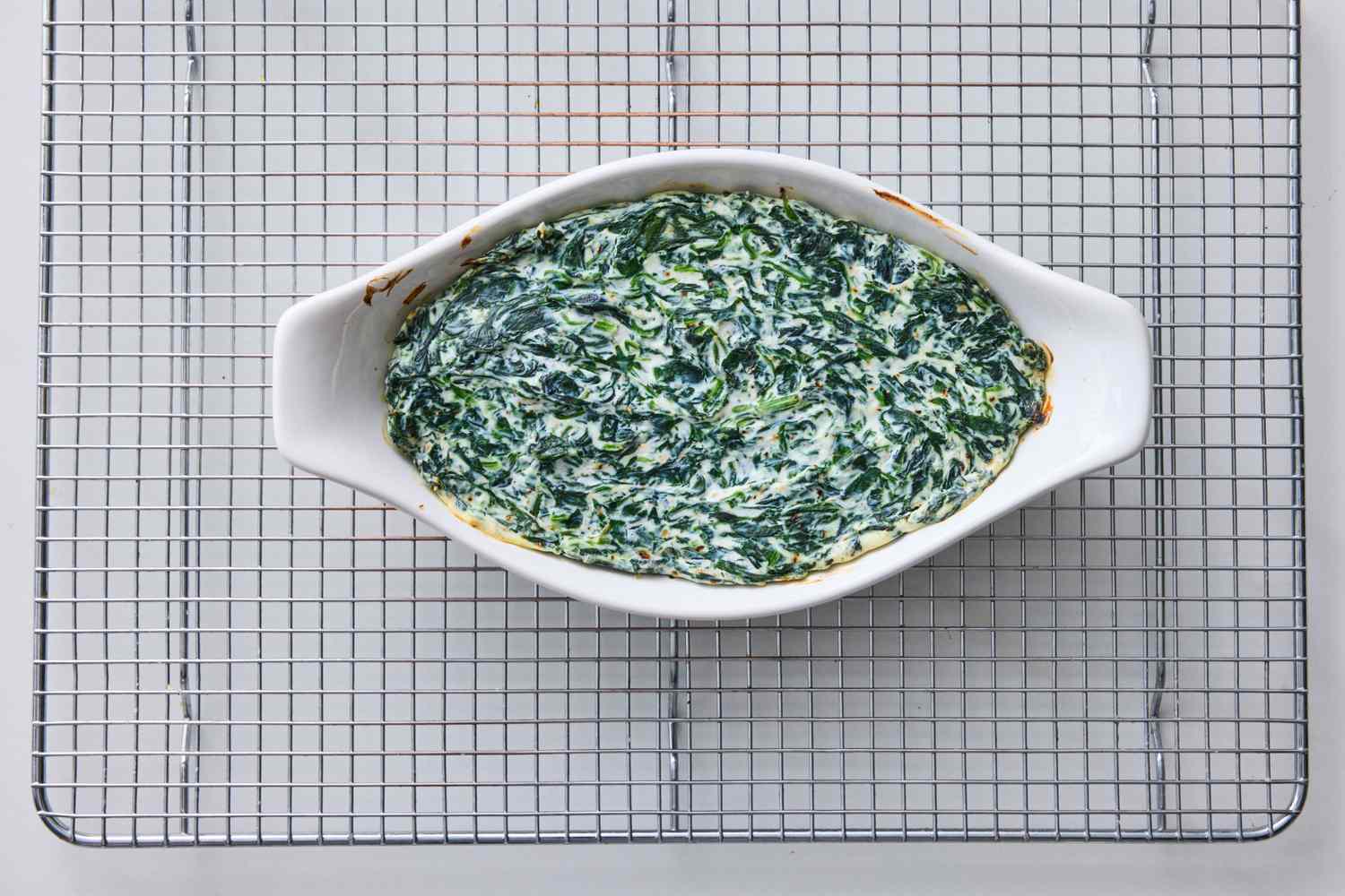 A serving of spinach dip in a white oval dish placed on a wire cooling rack