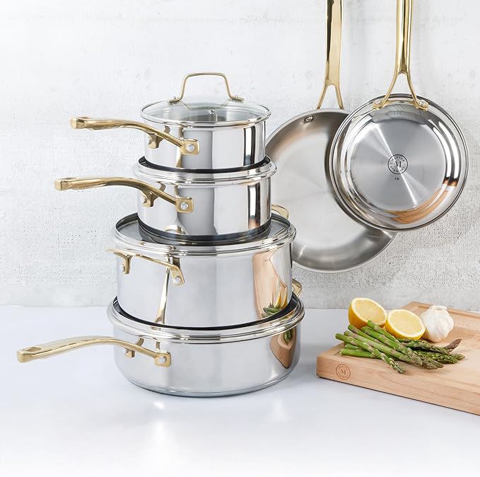 A set of stainless steel cookware including pots and frying pans with goldcolored handles displayed on a surface with some vegetables