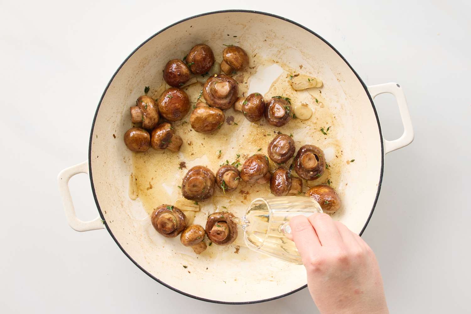 Sauted whole mushrooms with garlic and herbs in a white skillet a hand pouring liquid into the skillet