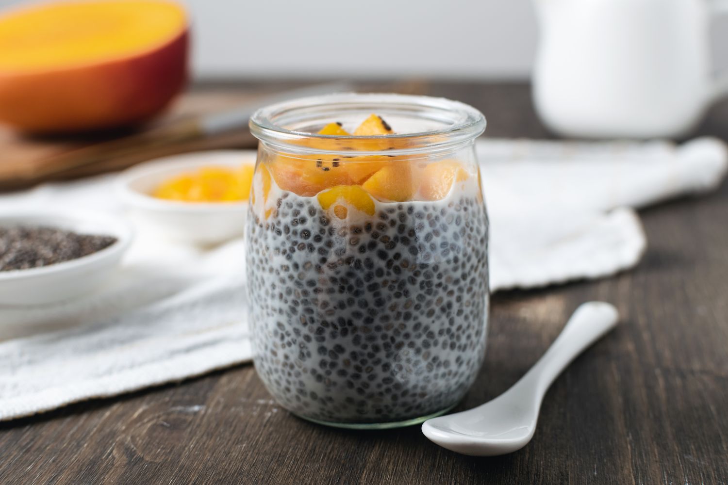 A jar of chia pudding topped with mango chunks on a wooden table with a ceramic spoon next to it
