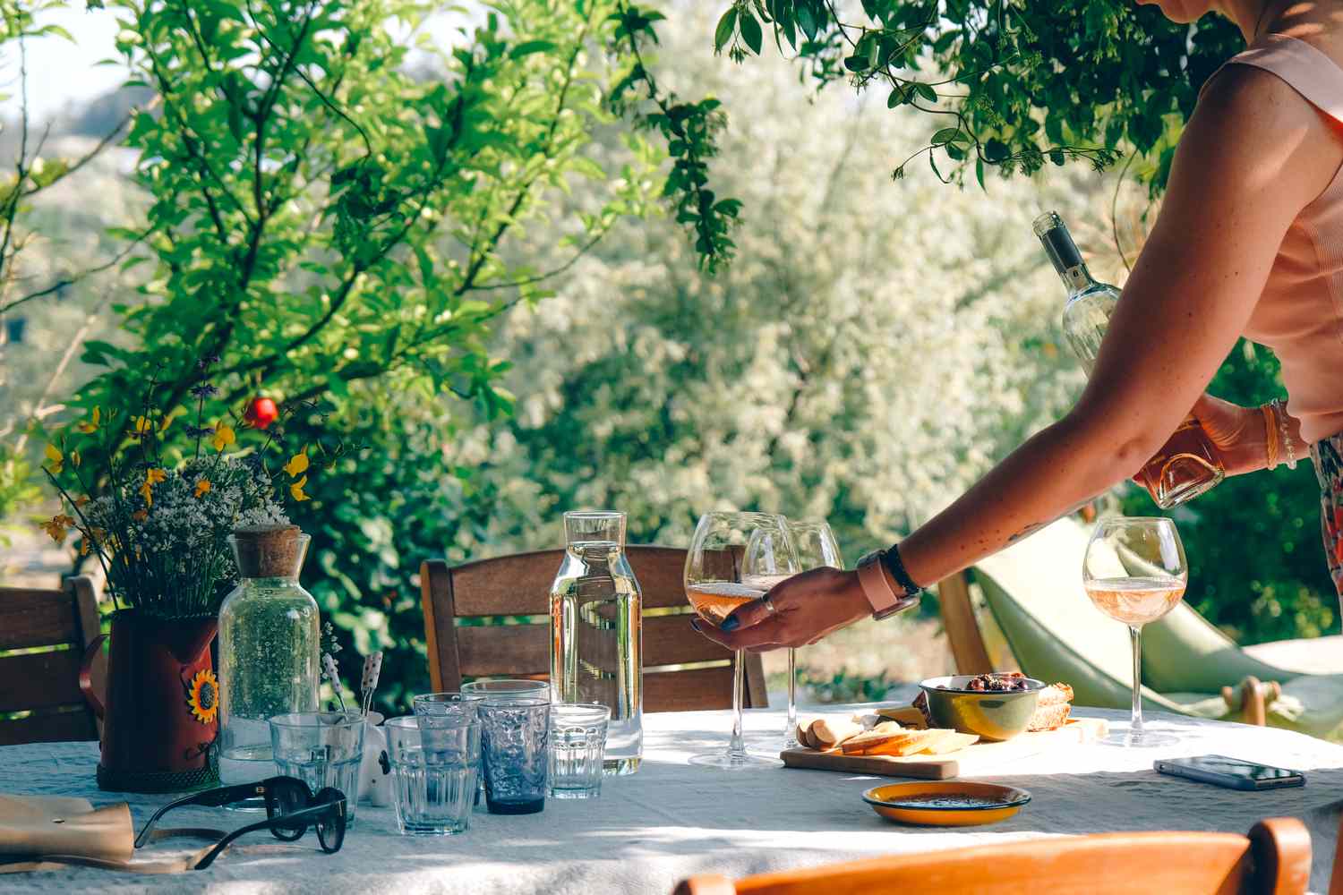 Table set outdoors with various drinks and dishes person pouring wine into a glass surrounded by greenery