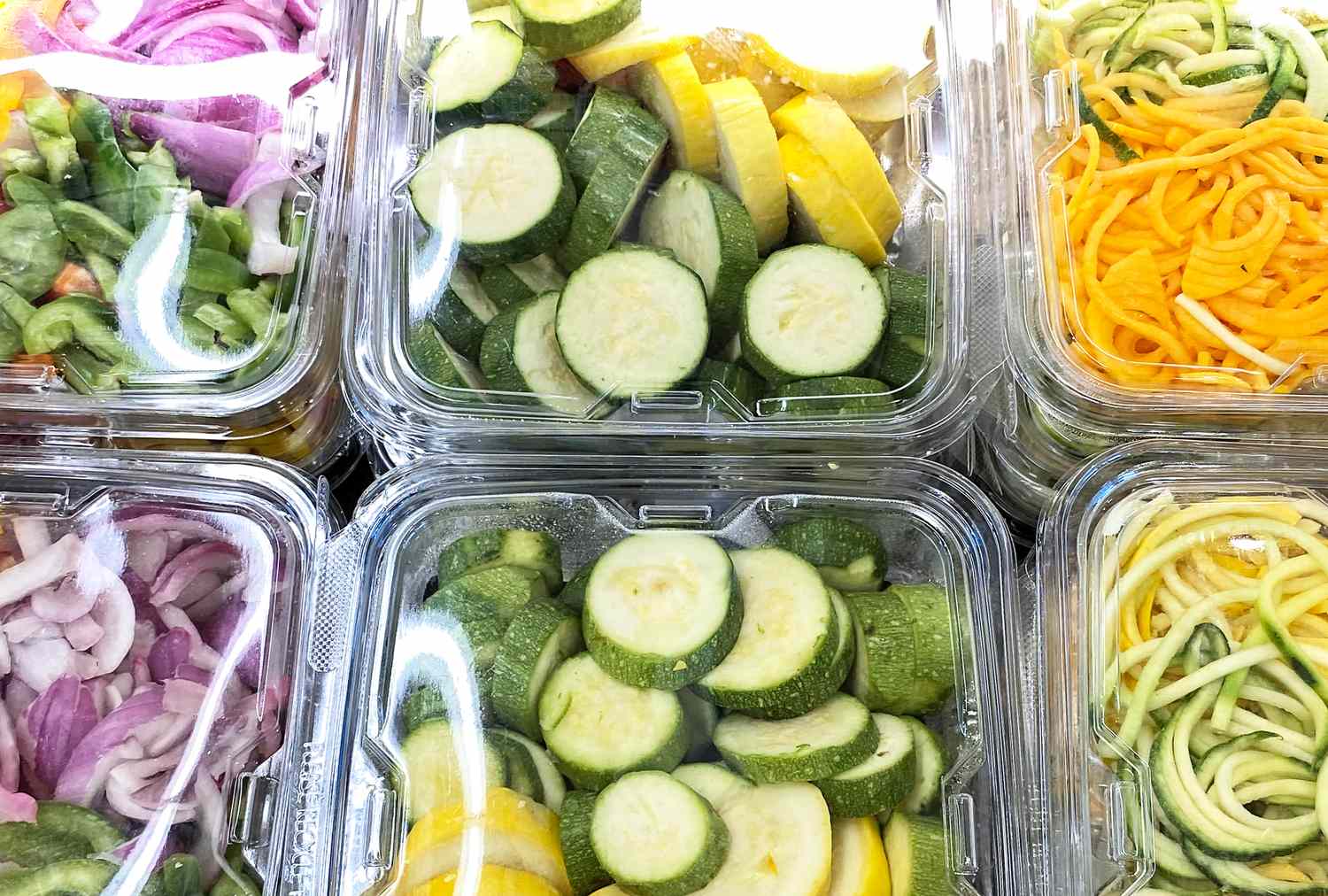 Assorted plastic containers filled with precut vegetables including zucchini slices and spiralized vegetables