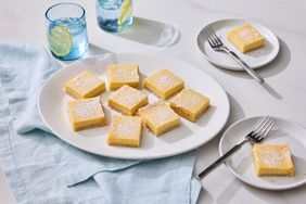 Creamy lemon bars with powdered sugar on a white plate served with drinks and utensils on the side