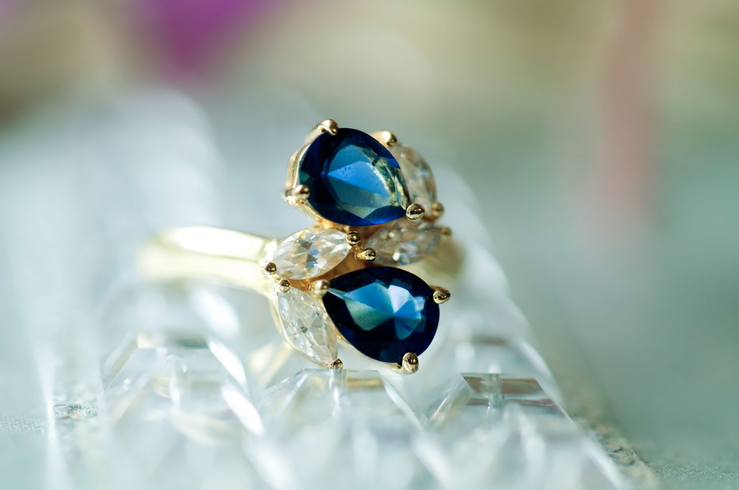 A gold ring with blue and clear gemstones displayed on a reflective surface