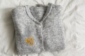 A folded gray cardigan with a visible yellow stain on the front lying on a white fabric surface