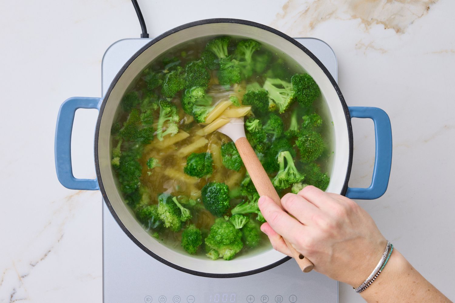 A pot containing broccoli and pasta being stirred by a hand with a wooden spoon