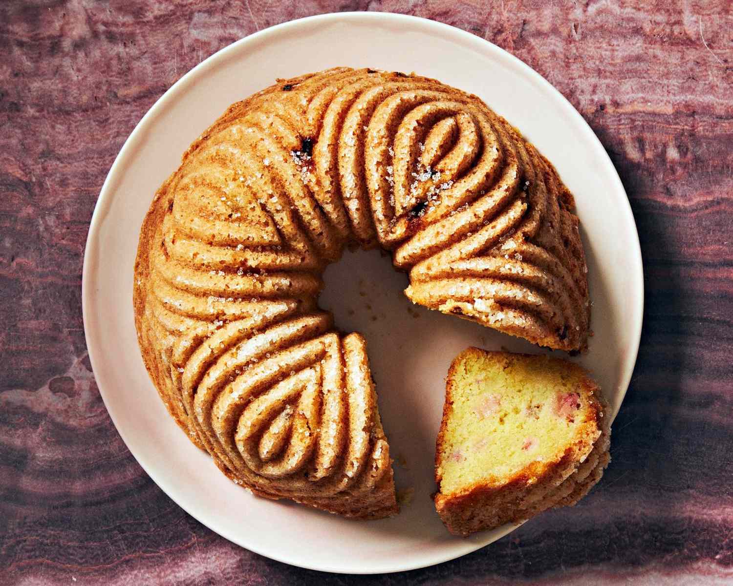 A olive-oil bundt cake on a white plate with a slice removed revealing a textured moist interior on a dark surface