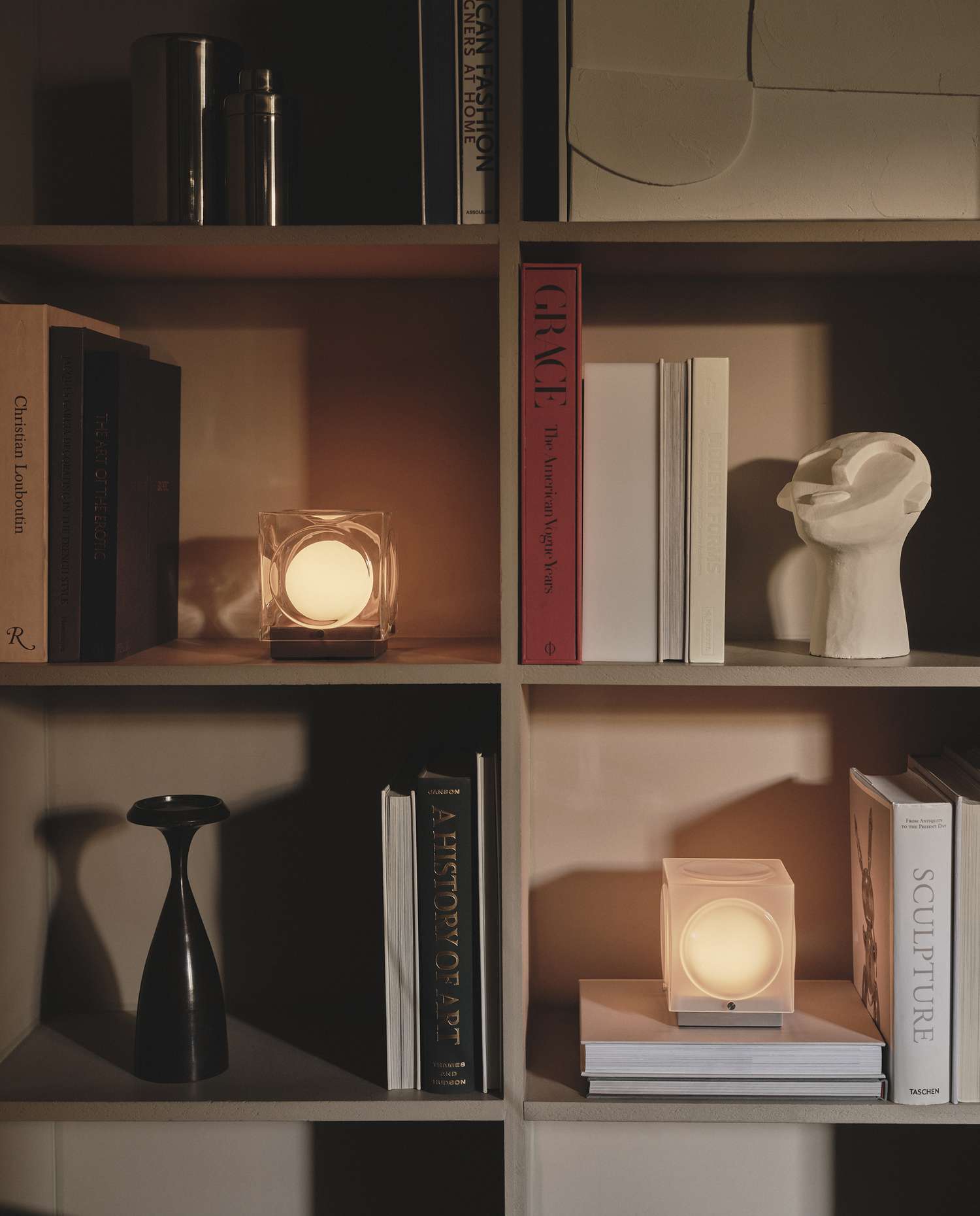 A shelf with books decorative objects and lit cubeshaped table lamps