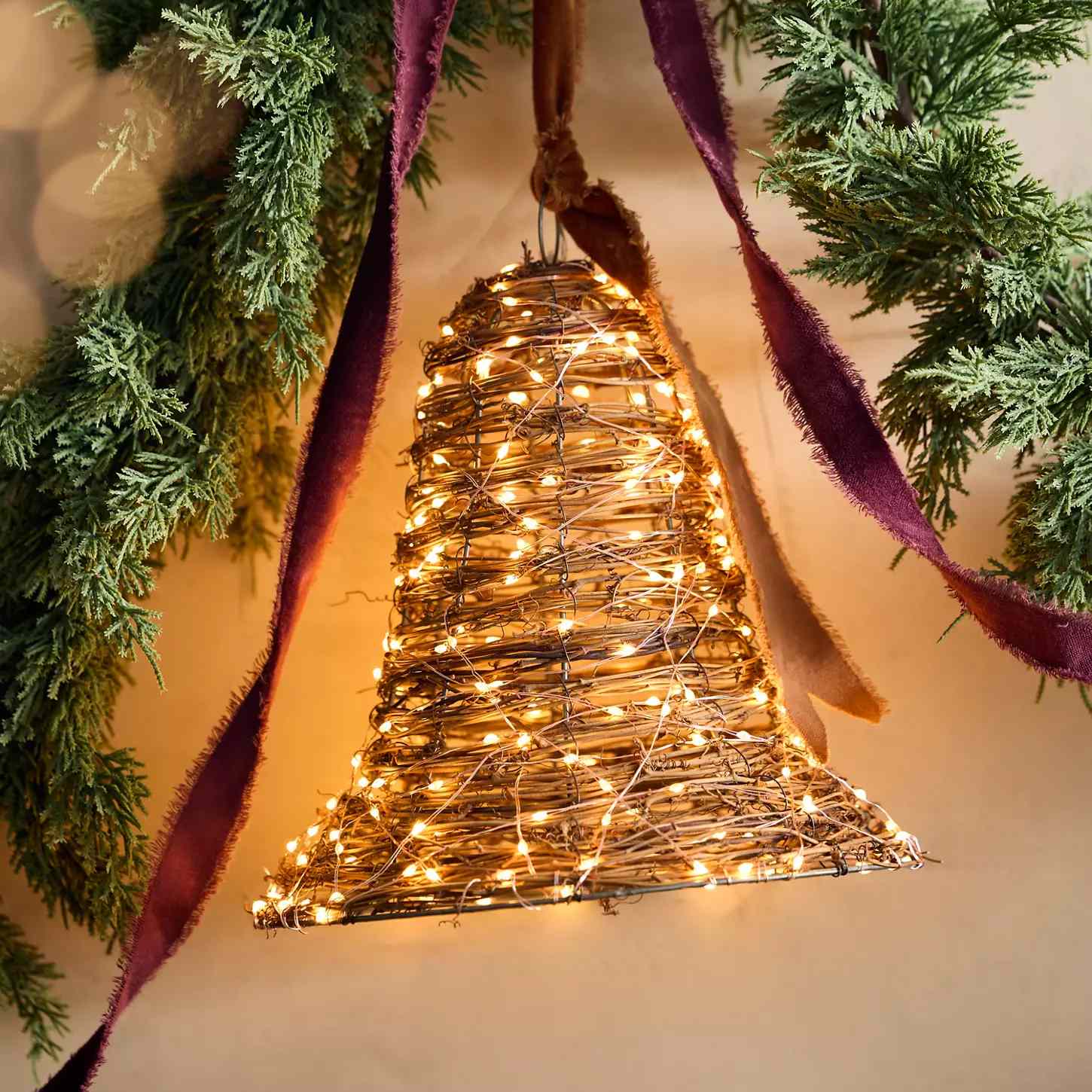 A decorative bell made of twigs and lights hung with a ribbon and surrounded by greenery