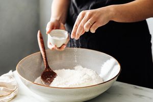 A person sprinkling an ingredient into a bowl of flour with a wooden spoon resting in the bowl on a table
