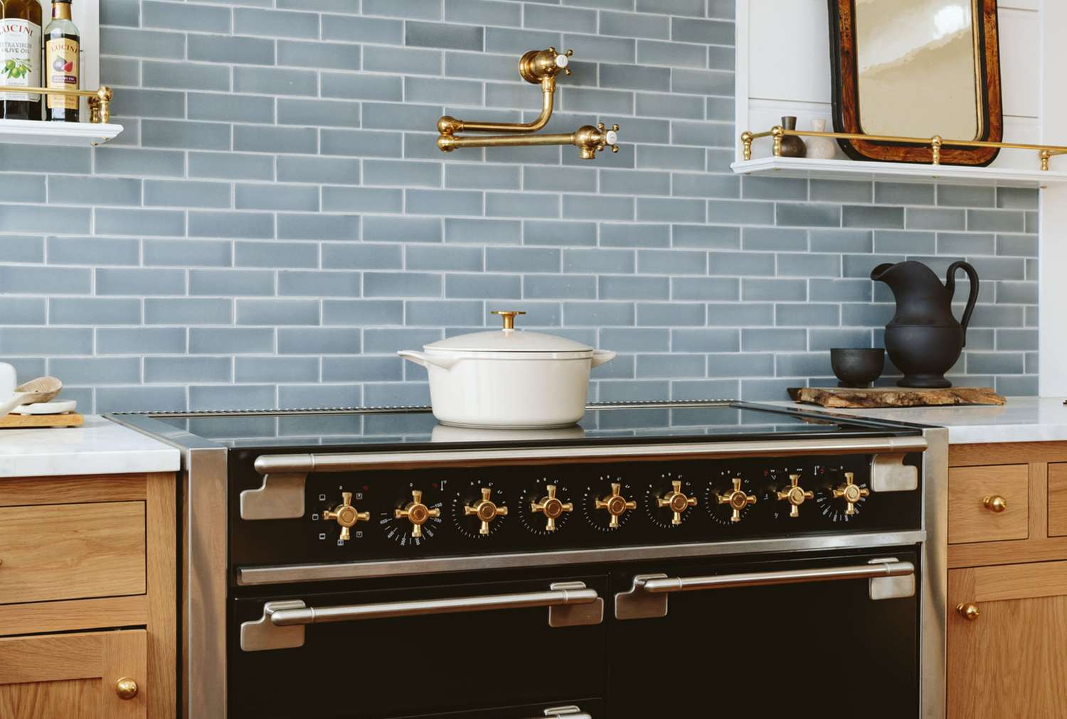 A black range oven with brass accents beneath a blue tiled backsplash a white pot on the stove and a pitcher on the counter nearby