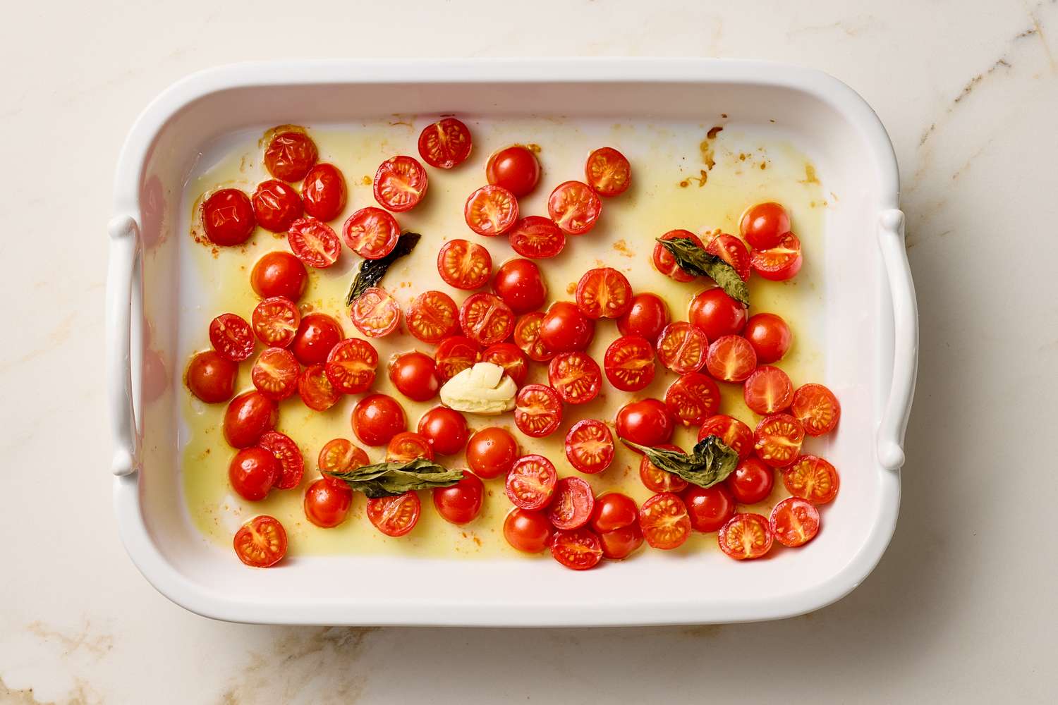 A baking dish with halved cherry tomatoes olive oil a garlic clove and fresh herbs before cooking