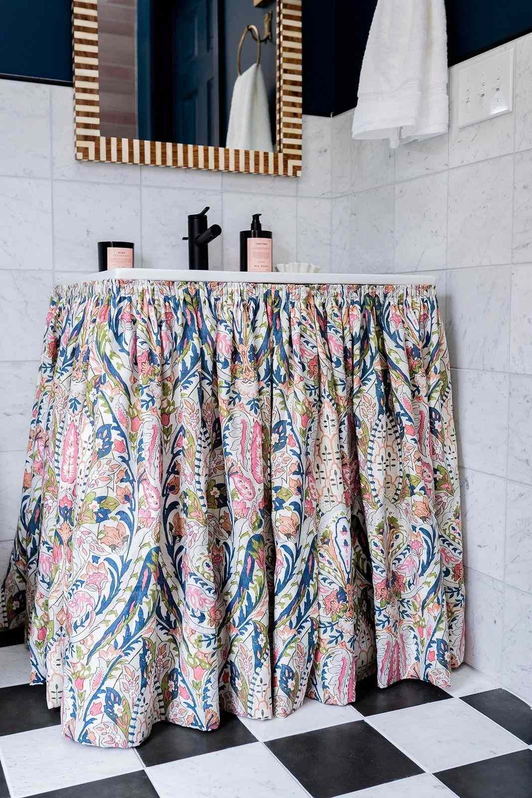 A bathroom sink with a patterned skirt covering the base a checkered floor and a striped frame mirror above