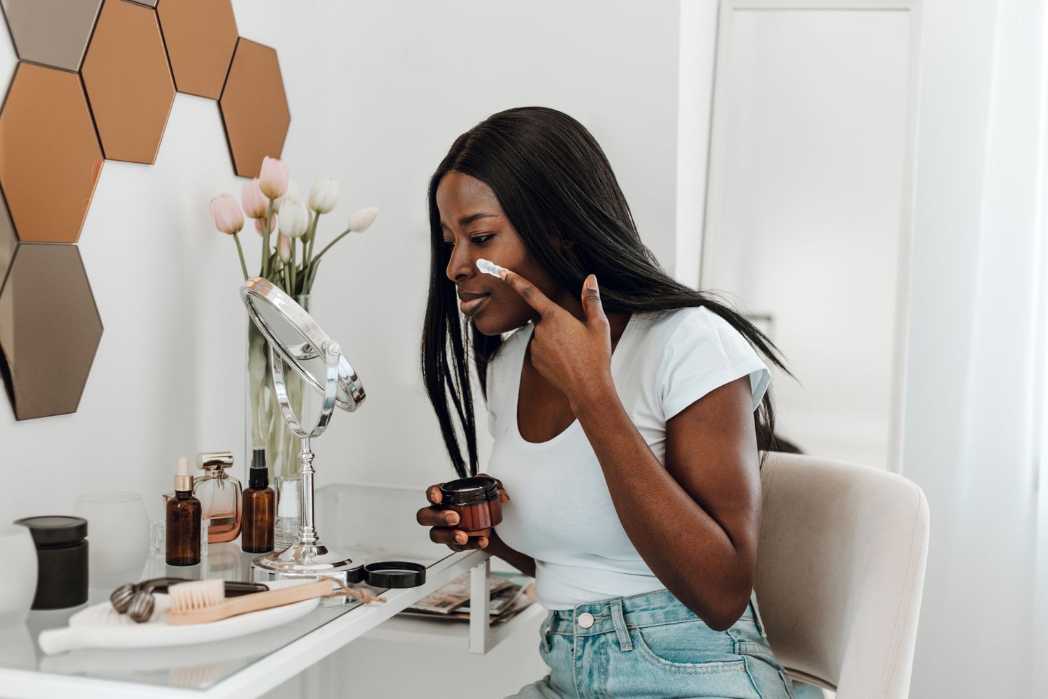A person applying skincare cream in front of a mirror at a table with cosmetic items a decorated hexagonal wall mirror is in the background