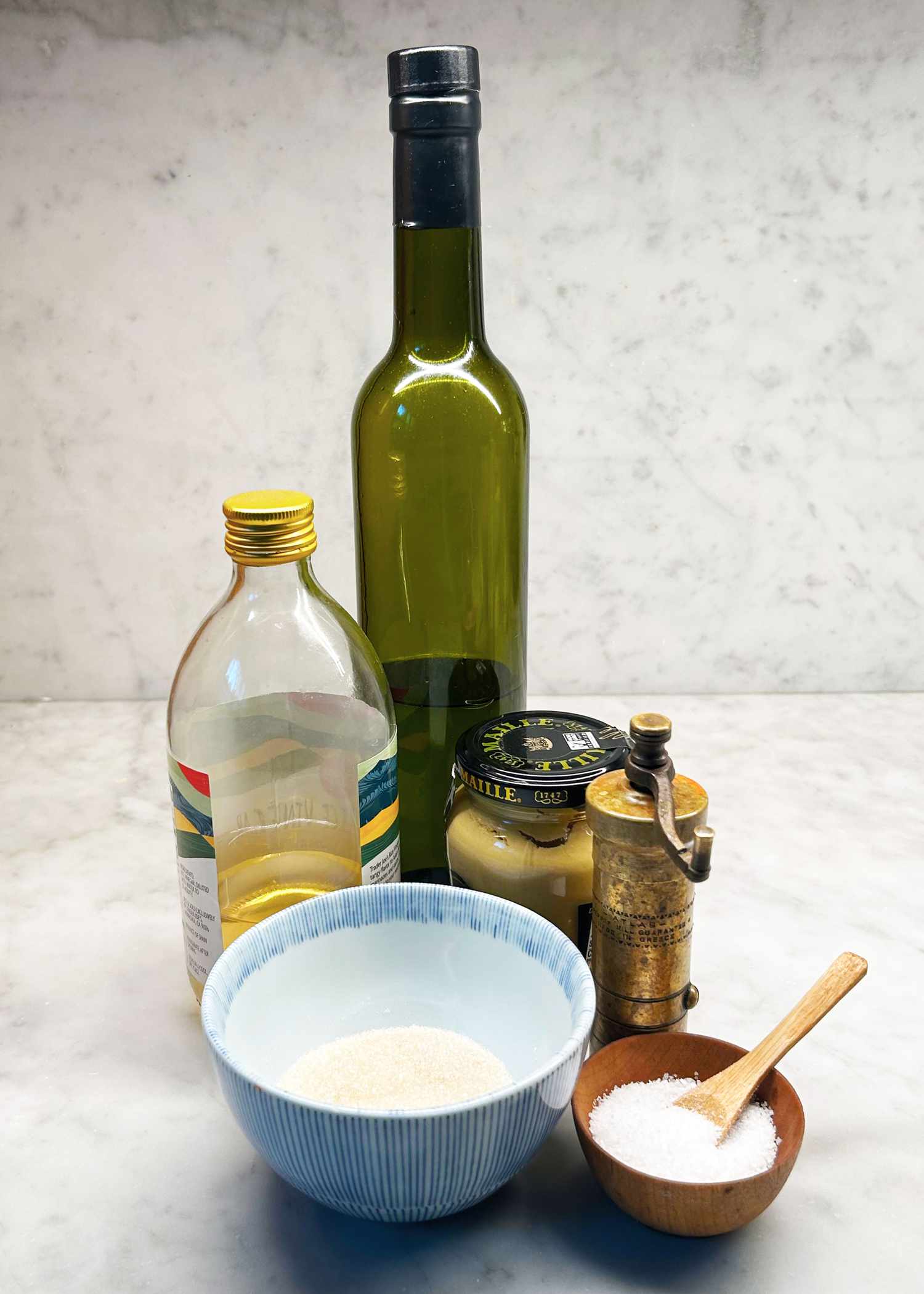 Ingredients for making vinaigrette, including olive oil, mustard, vinegar, sugar, salt, and pepper grinder, arranged on a counter