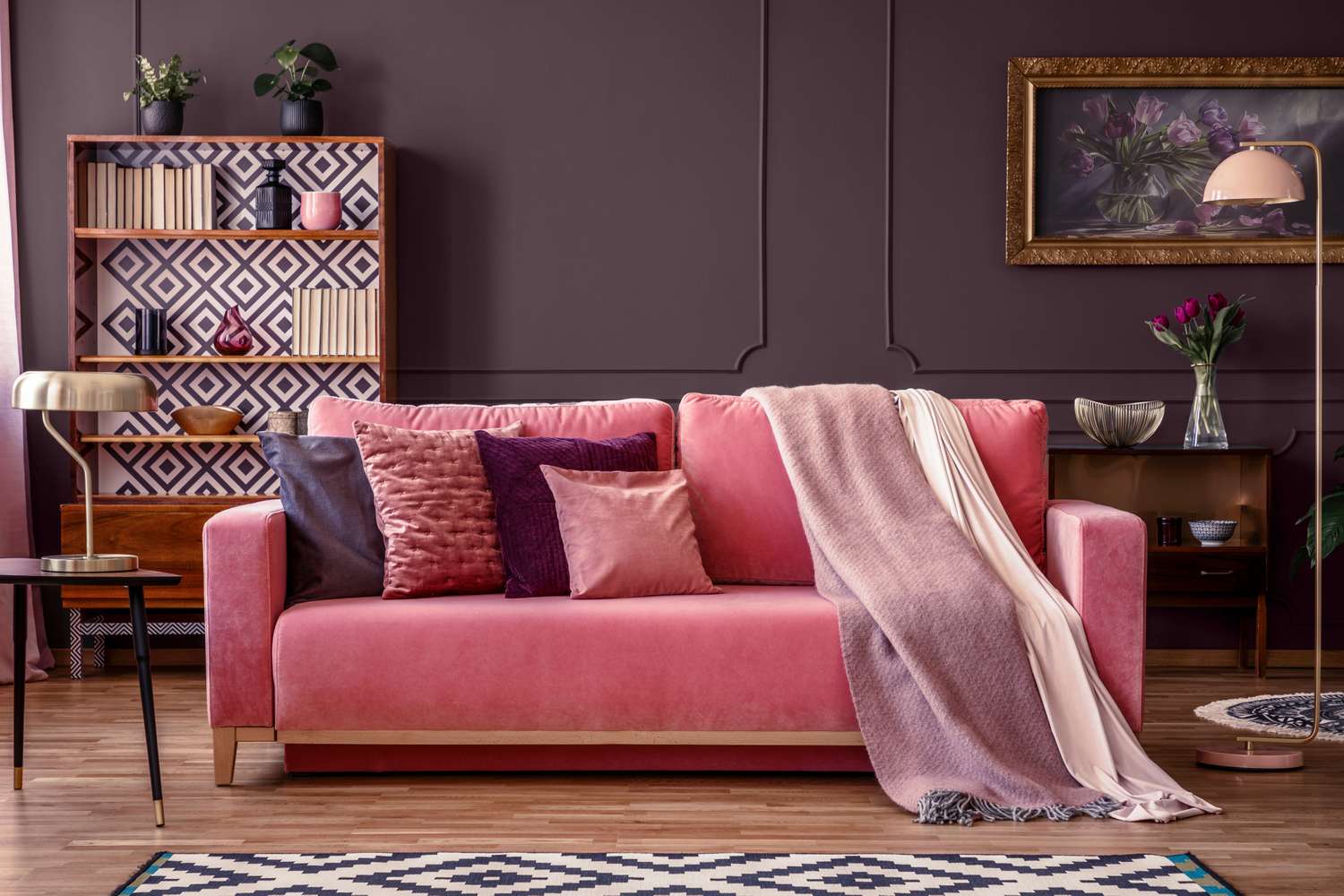 A living room with a sofa adorned with cushions and a throw blanket bookshelf and decor framed picture and a side table with a lamp