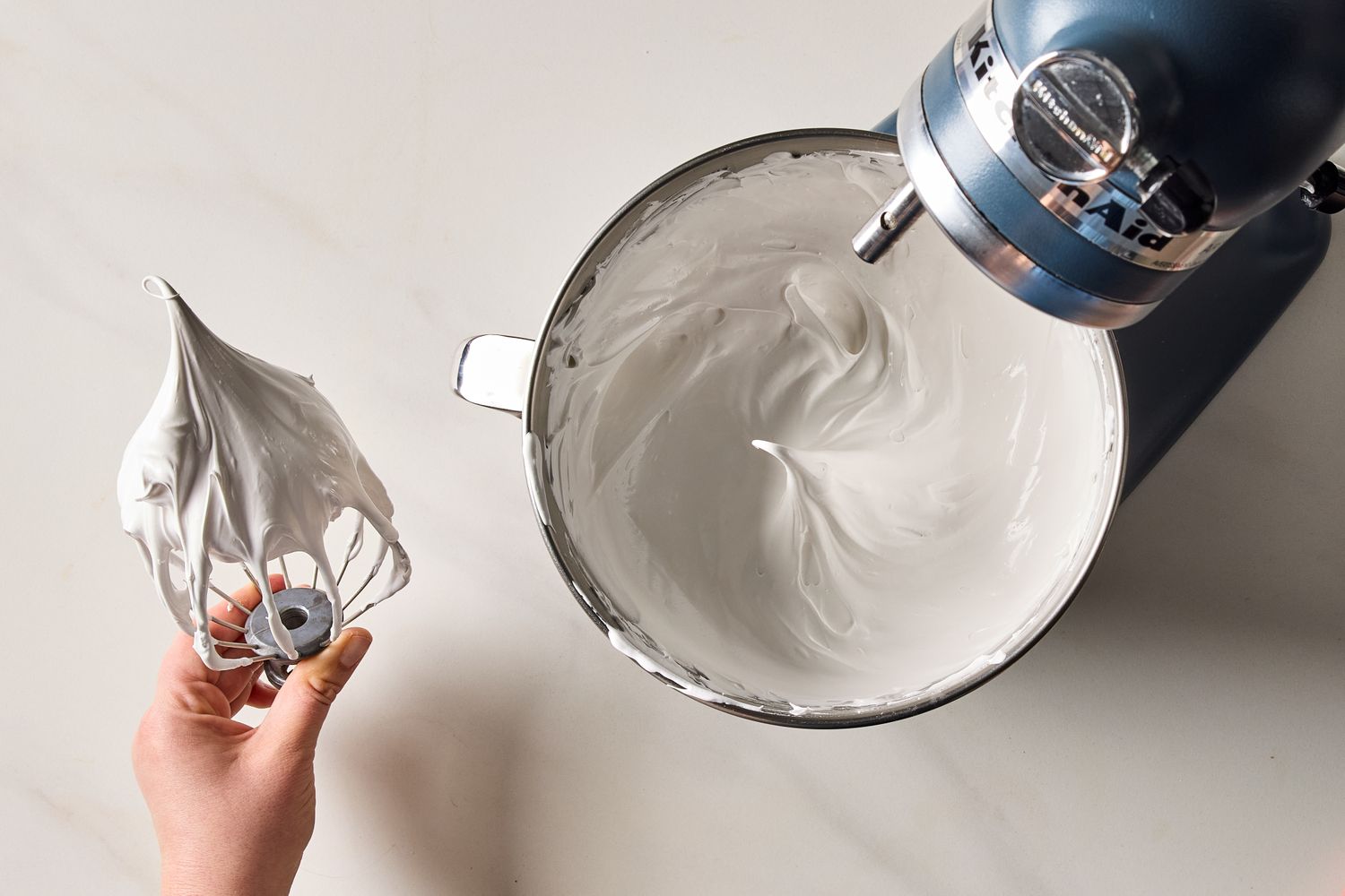 A bowl of prepared marshmallow frosting on a stand mixer with a hand holding a whisk attachment covered in frosting