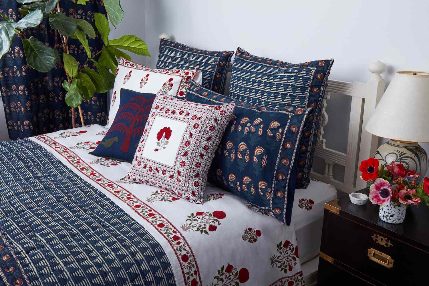A bed with a floral patterned bedspread multiple decorative pillows and a bedside table with a lamp and flowers