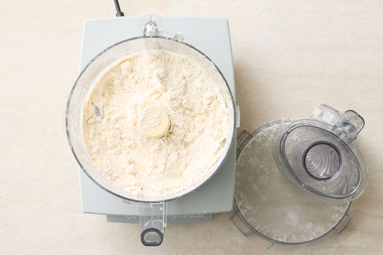 Food processor bowl with flour and blade lid placed beside on counter