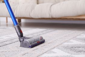 A vacuum cleaner being used on a carpet in a living room setting sofa in the background
