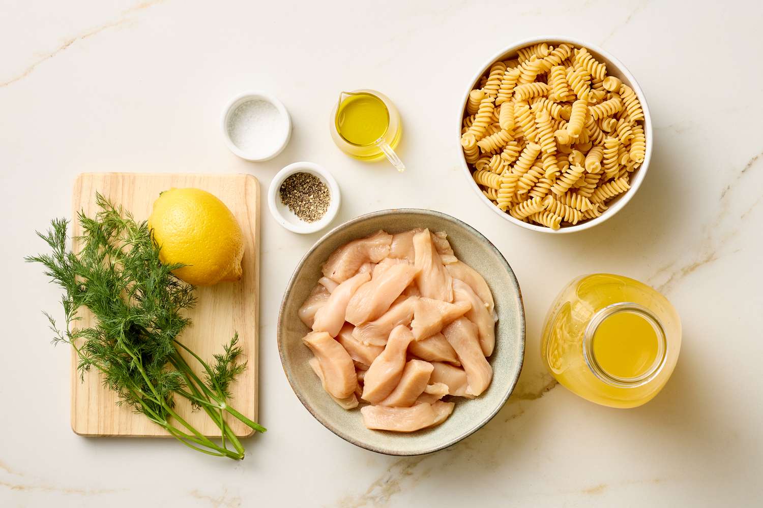 Ingredients for a pasta dish including uncooked rotini raw chicken pieces lemon dill and seasonings