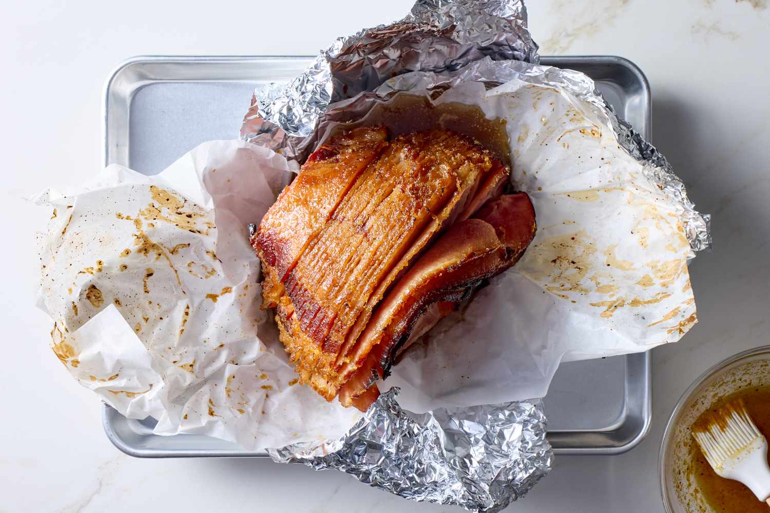 A cooked spiralcut ham partially wrapped in foil and parchment paper on a baking sheet