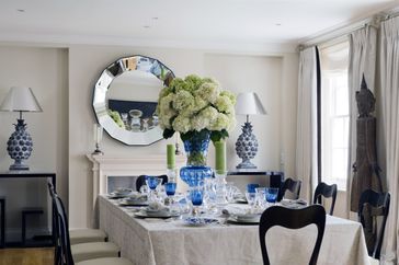 A formal dining table with a centerpiece of flowers and table settings a round mirror above a mantel in the background