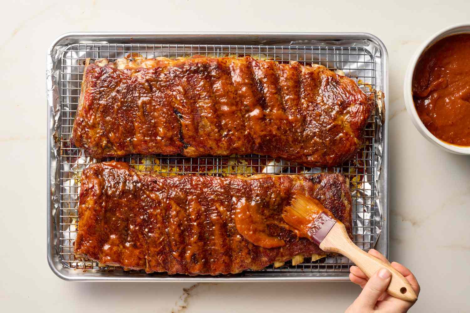 Cooked ribs on a baking sheet being brushed with barbecue sauce near a bowl of sauce