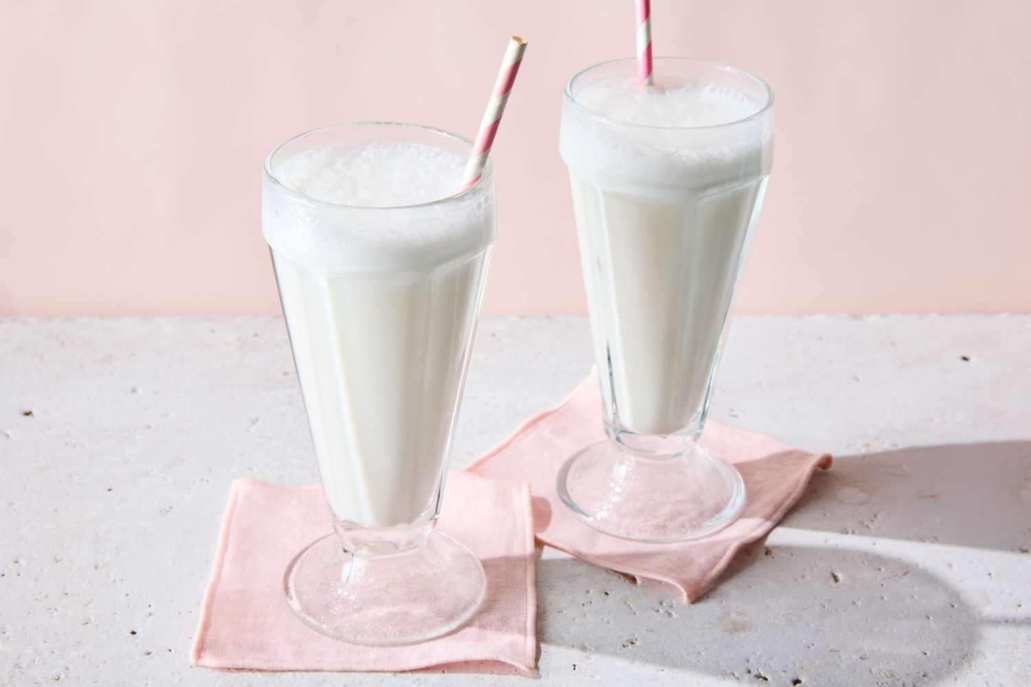 Two glasses of vanilla egg cream with straws on pink napkins
