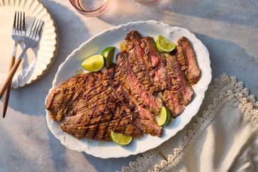 A white plate with sliced grilled flank steak garnished with lime wedges, placed on a table