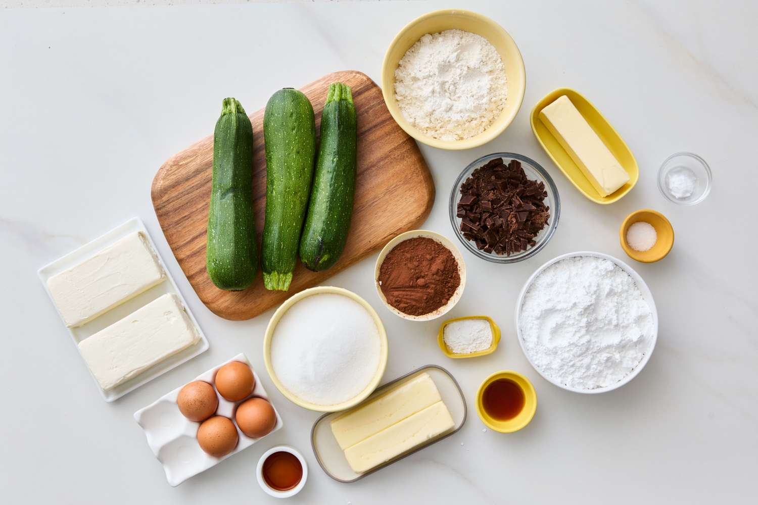 Ingredients for a zucchini sheet cake including zucchinis eggs butter sugar cocoa powder flour and cream cheese laid out on a countertop