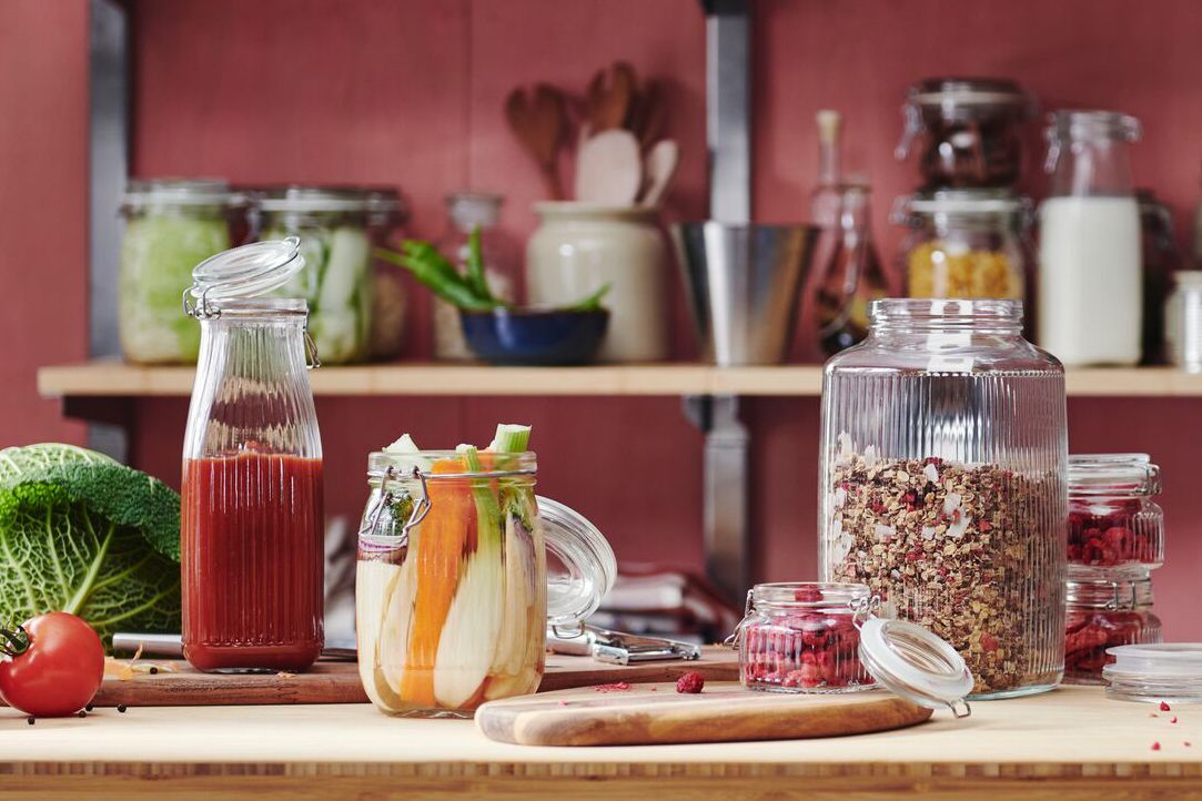 A countertop with jars of food including grains vegetables and juice with a blurred kitchen background featuring shelves and utensils