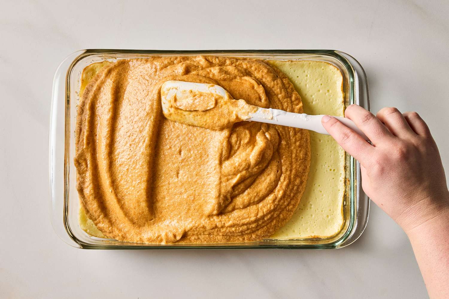 A hand spreading pumpkin mousse onto cheesecake in a rectangular glass dish with a spatula