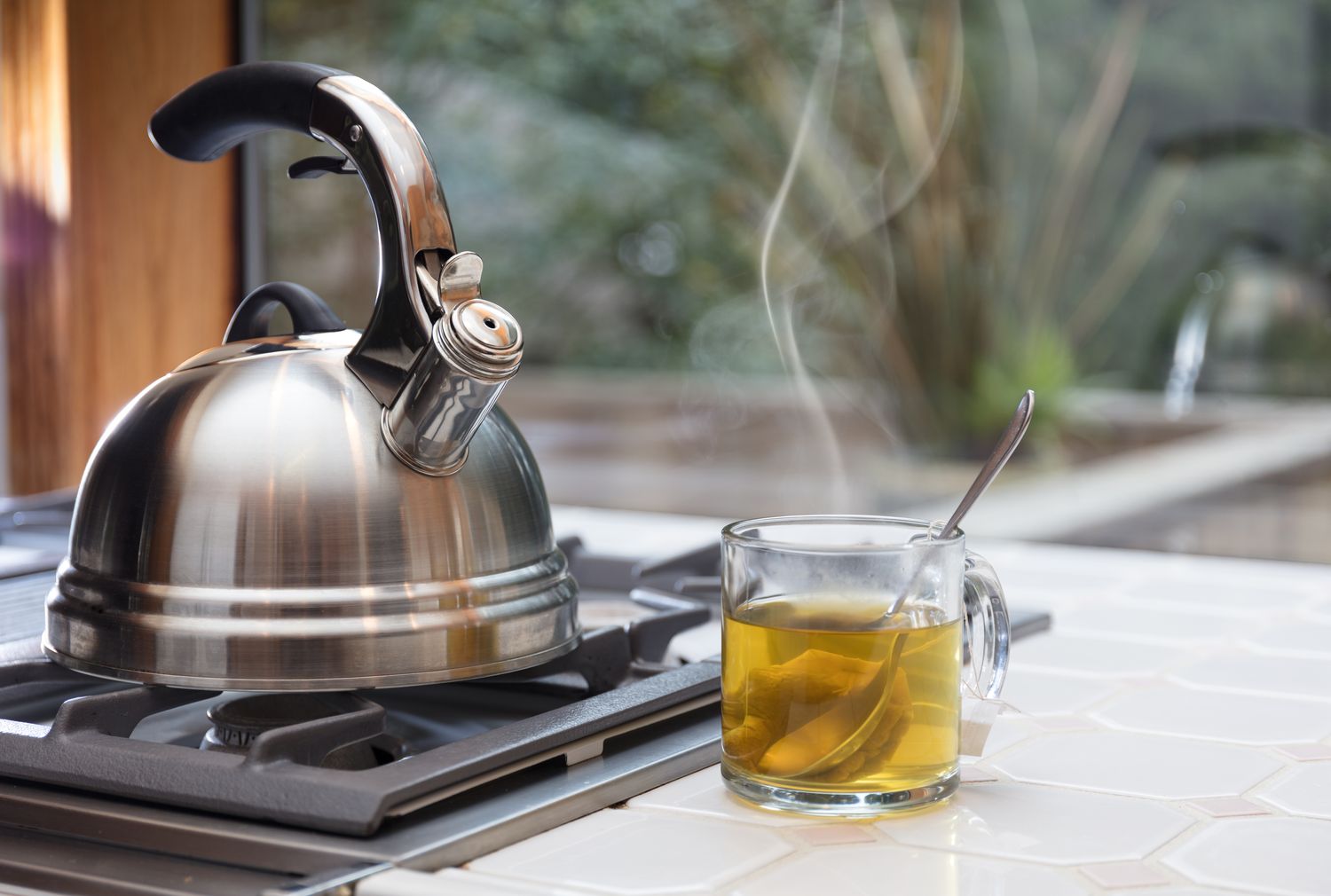 A stovetop kettle on a burner next to a steaming cup of tea on a tiled surface