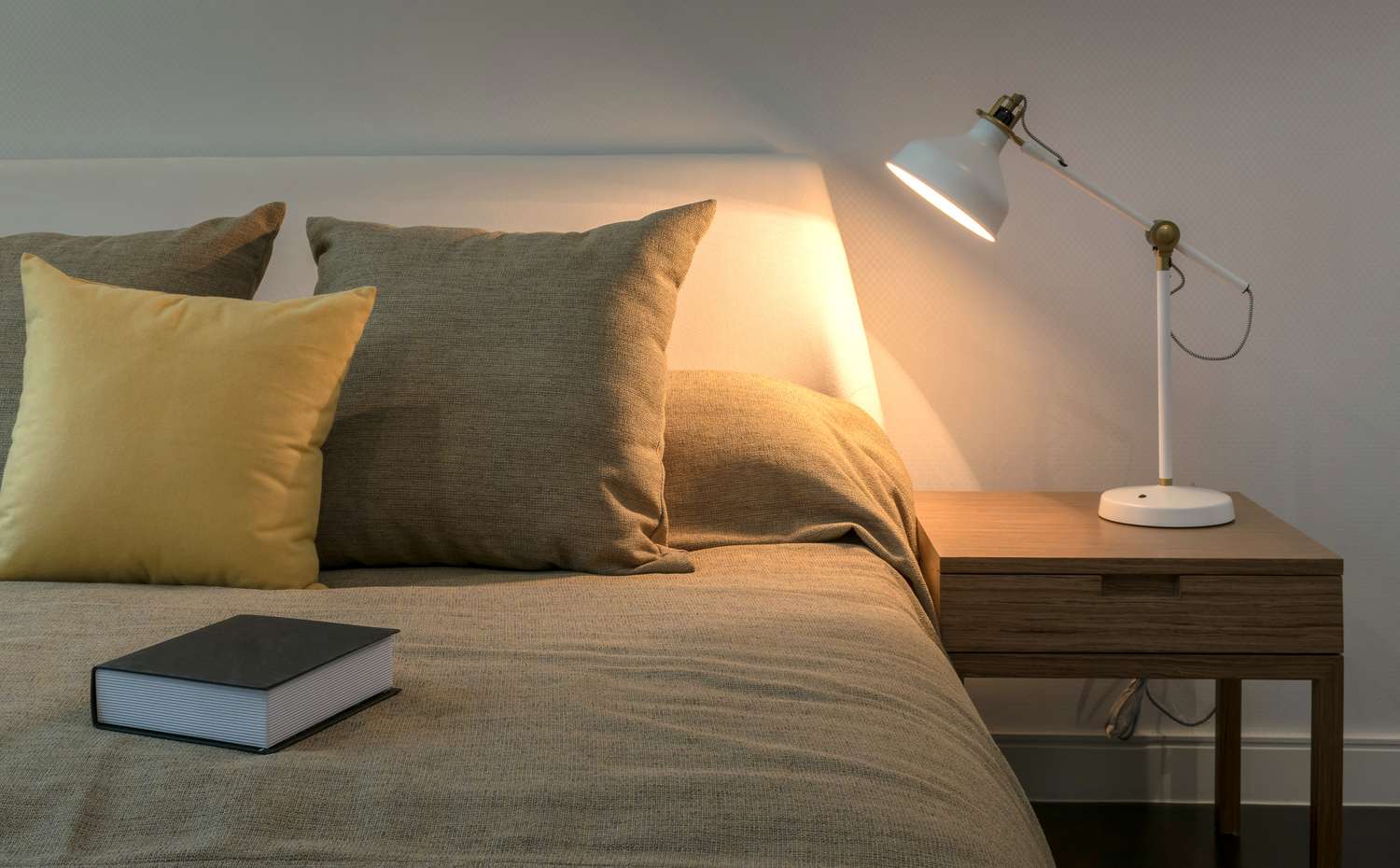 A bed with pillows a wooden nightstand a lamp and a book