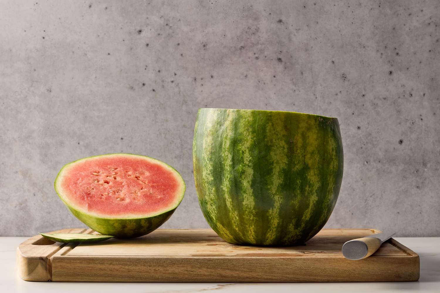 Halved watermelon on a wooden cutting board with an empty carved bowl and knife on a neutral background
