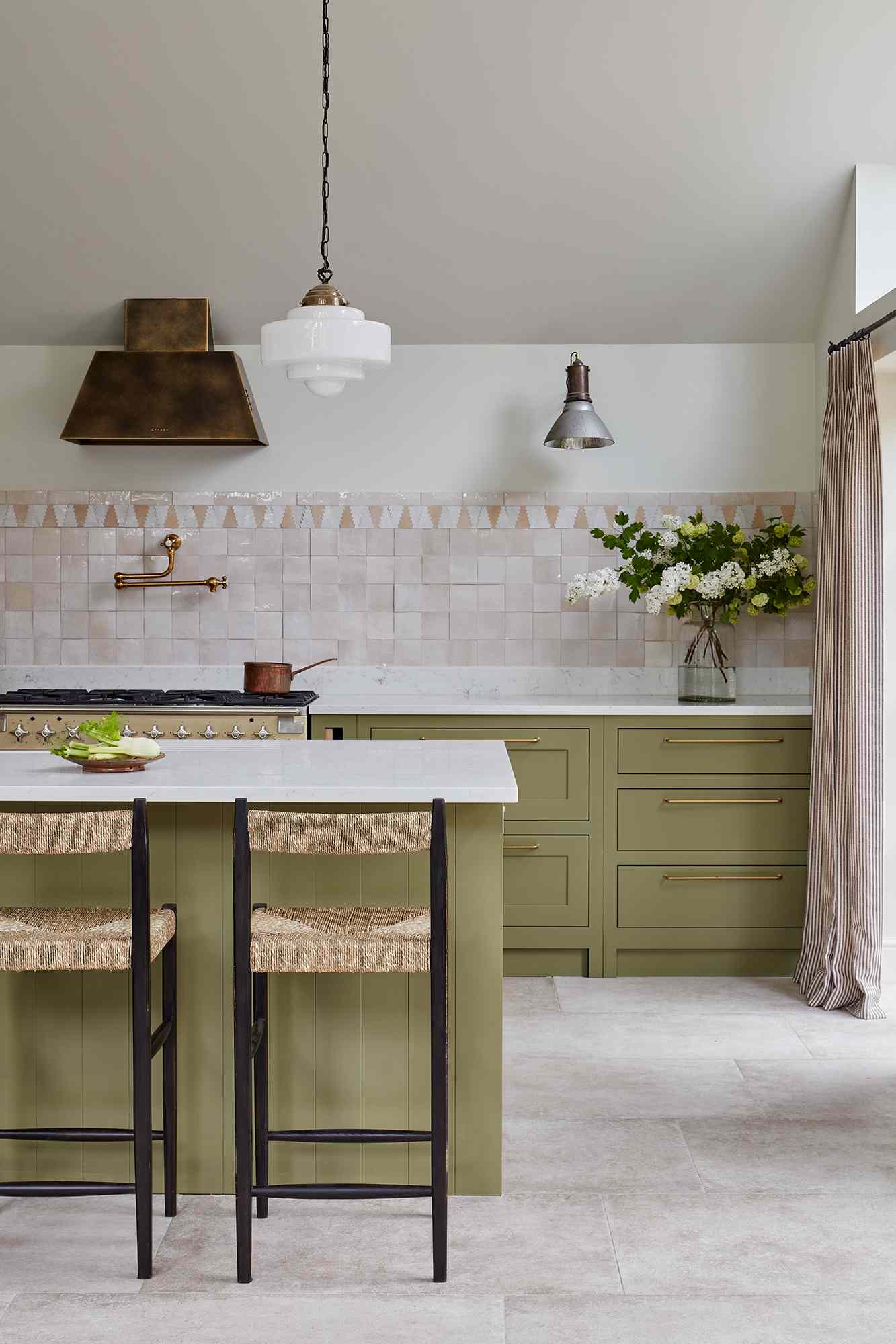 Kitchen design by Camilla Masi of Otto Tiles: muted apple green cabinetry, brass hardware, woven barstools, and a soft pink zellige tile backsplash. A statement bronze range hood and vintage-inspired lighting add warmth and character.