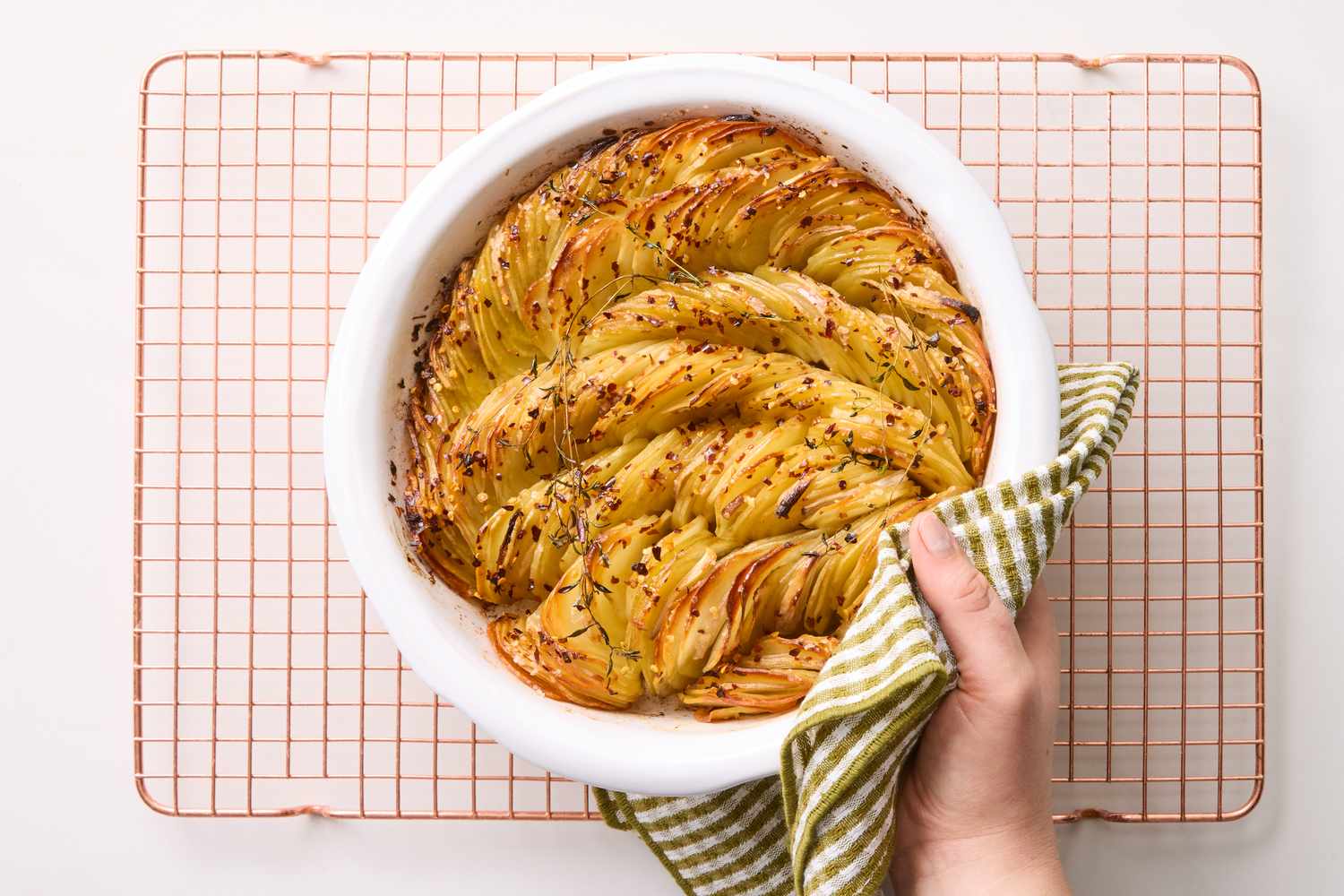 A baked potato dish arranged in layers in a white dish shown on a wire rack with a hand holding it using a kitchen towel