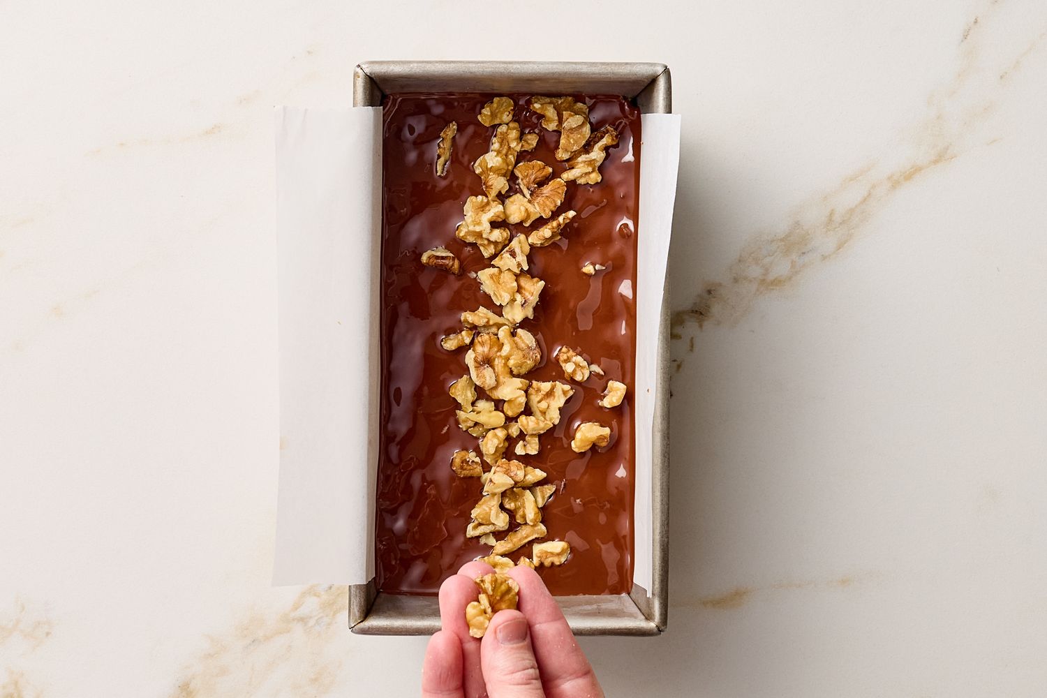 Someone adding walnut pieces to a tray of dark chocolate mixture