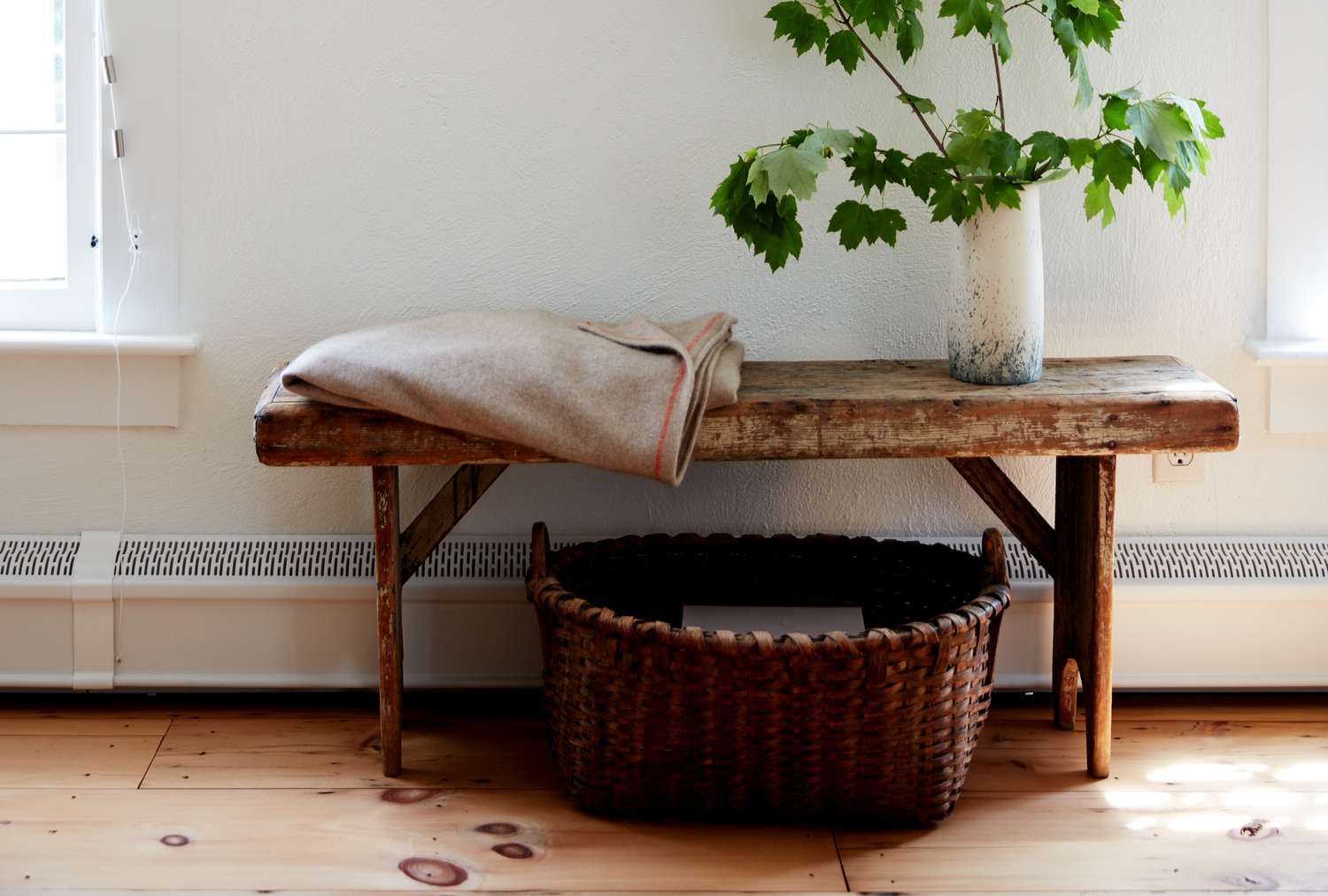 Rustic wooden bench with a blanket and a vase with branches a wicker basket underneath located by a light windowed wall