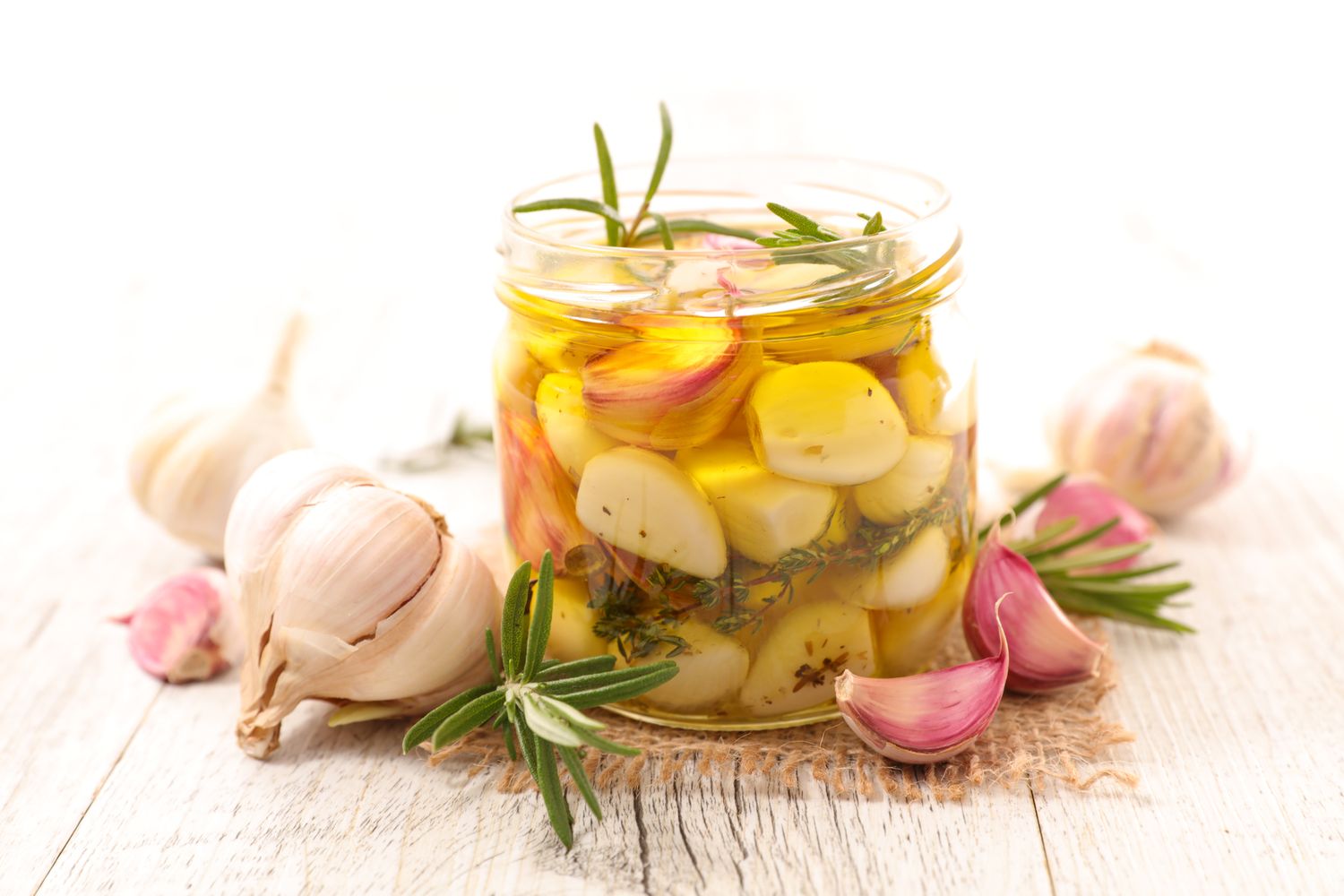 A jar of preserved garlic cloves in oil with herbs surrounded by fresh garlic bulb and cloves on a rustic white surface