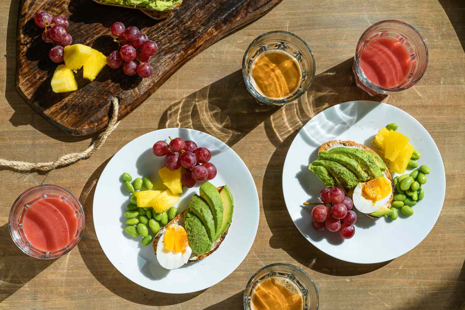 Two plates with avocado halfboiled egg grapes pineapple edamame and served with glasses of juice and coffee on a table