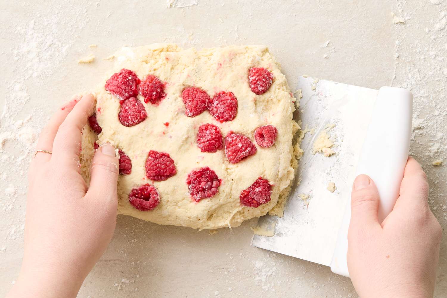 Hands shaping dough with raspberries using a pastry scraper