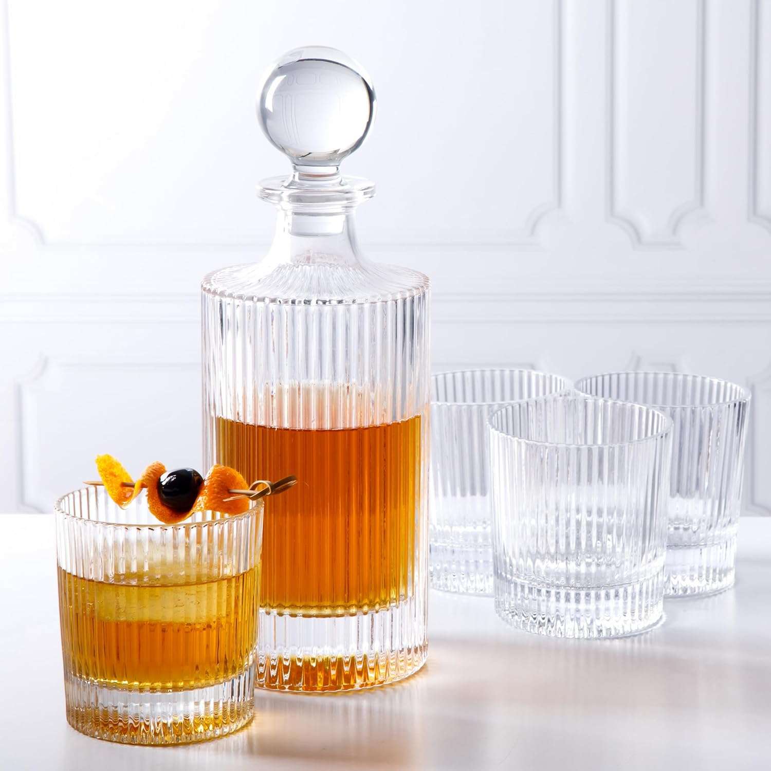 A crystal decanter with whiskey and a garnished glass accompanied by additional empty glasses placed on a white surface