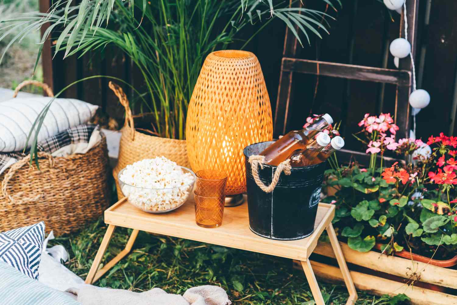 A cozy outdoor setup including a small table with a lamp popcorn glass and bucket of drinks surrounded by plants and cushions