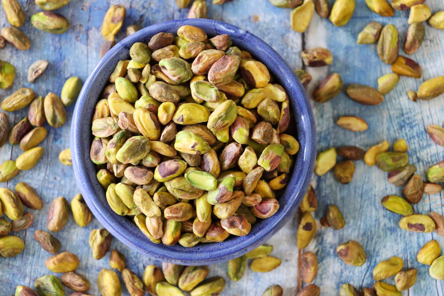 shelled pistachios in a blue bowl with more nuts on the surface