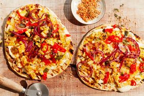 Two grilled pizzas topped with various vegetables and spices, with a pizza cutter and bowl of seasoning on a wooden surface