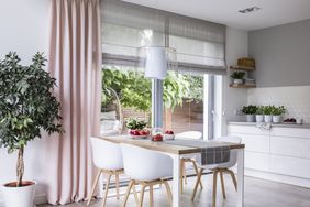 Gray roman shades and a pink curtain on big, glass windows in a modern kitchen and dining room interior with a wooden table and white chairs